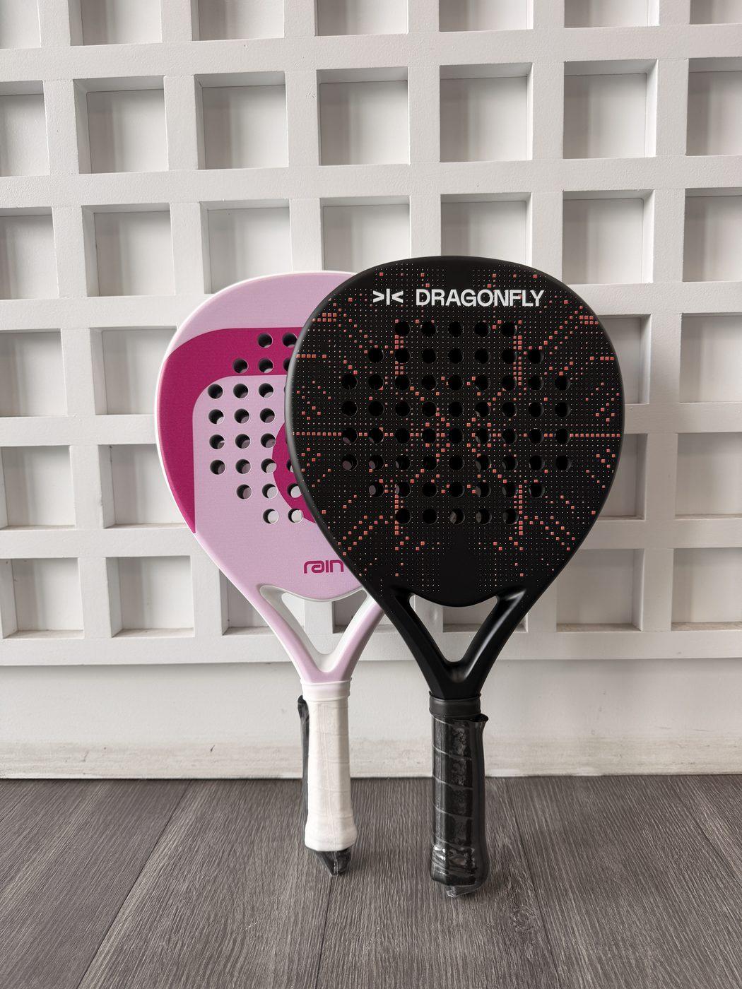 Custom pink 'rain' and black 'Dragonfly' padel rackets leaning against a white lattice wall
