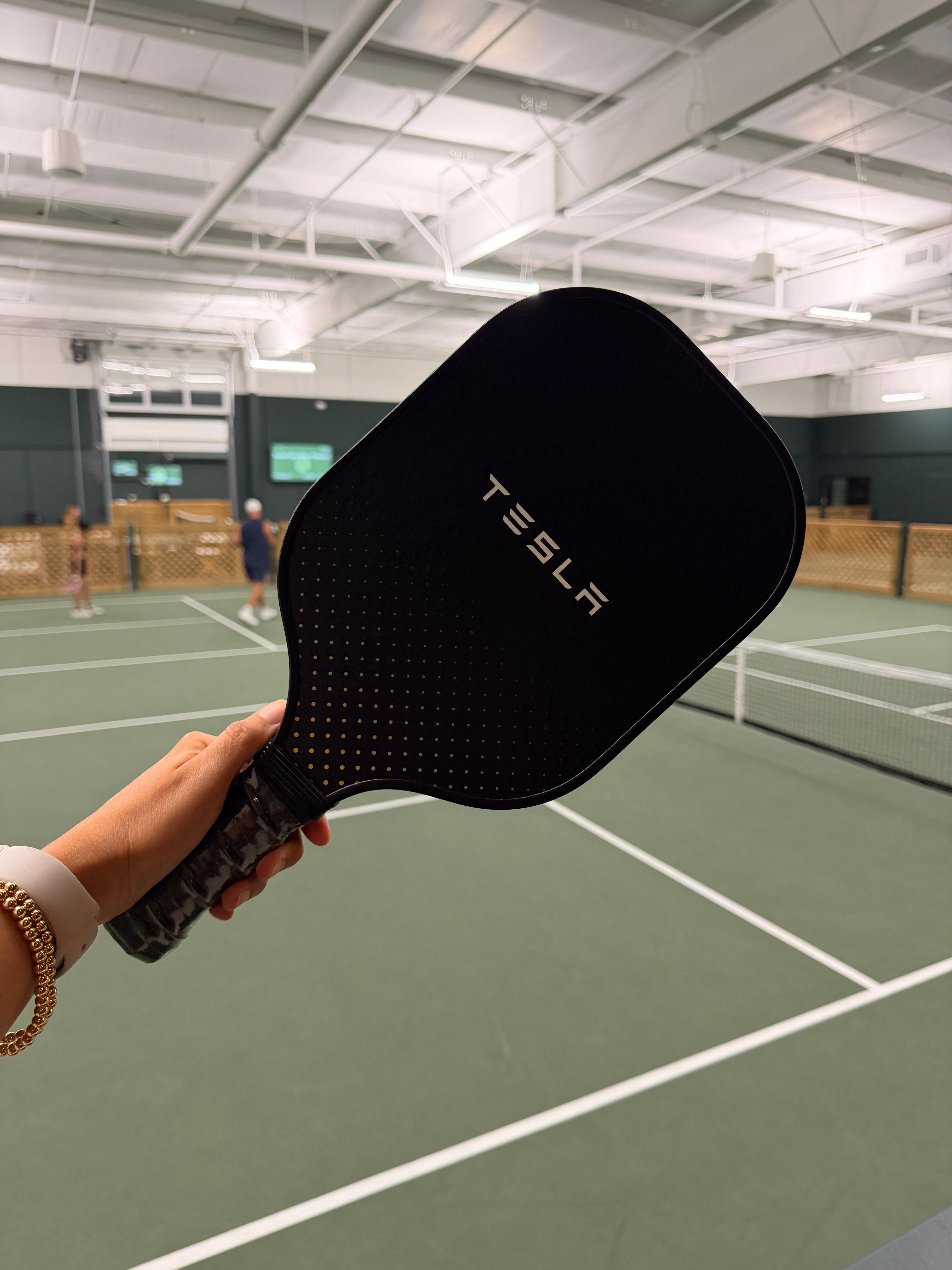 Custom Tesla carbon fiber pickleball paddle with a centered logo, on an indoor court