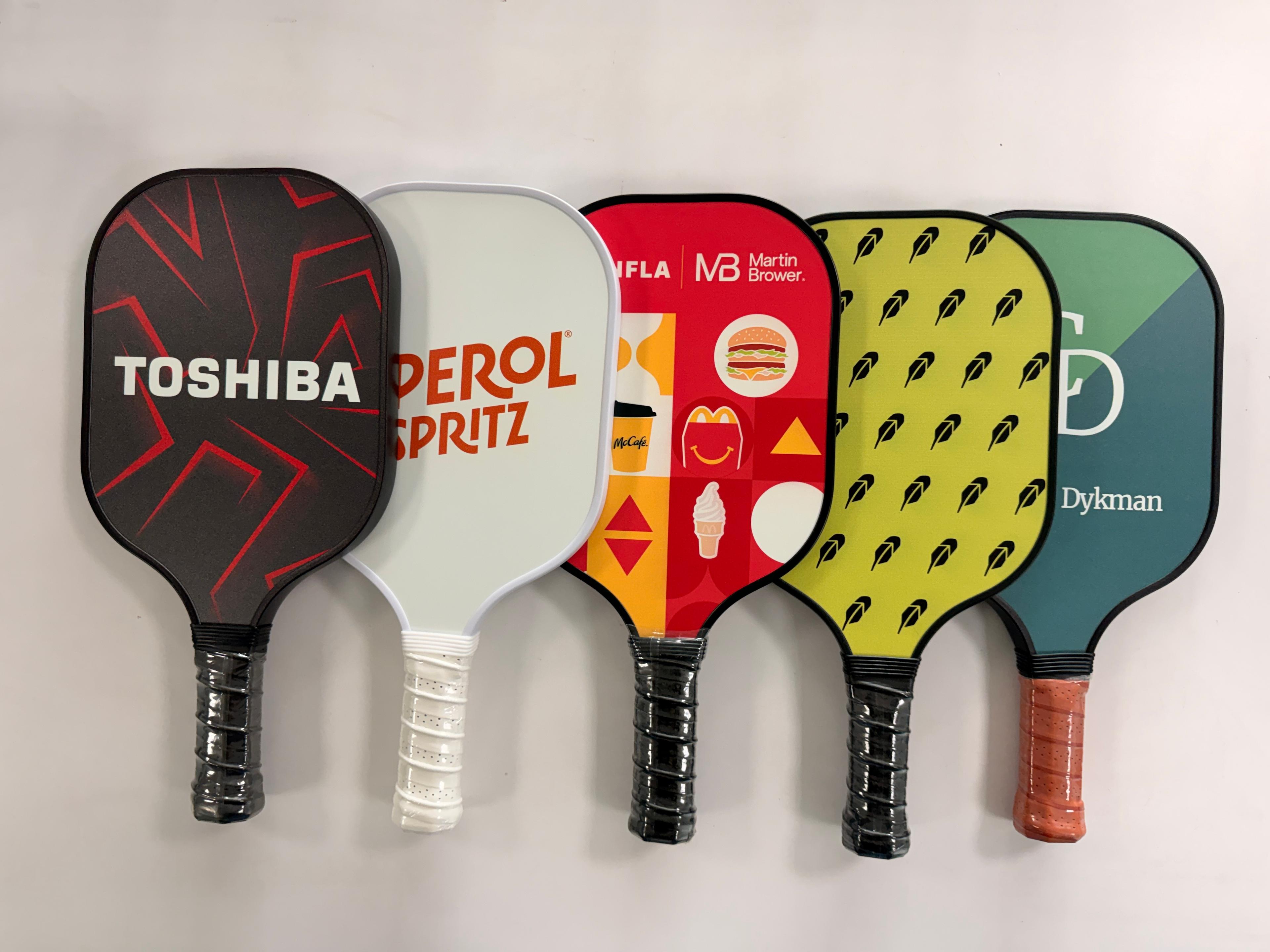 A lineup of custom-printed pickleball paddles — Toshiba, Aperol Spritz, McDonald's and more
